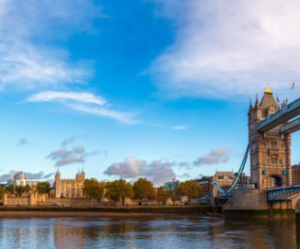 A picturesque view of Tower Bridge on the right and the Tower of London on the left, with a clear blue sky above. The River Thames is calm, reflecting the vibrant sky and historical architecture.