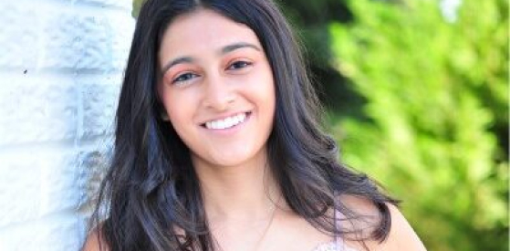 A person with long dark hair smiles while leaning against a white brick wall. They are outdoors with lush green foliage in the background.