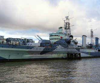 A large gray warship, HMS Belfast, is docked by the riverbank under a cloudy sky, with buildings in the background. The ship features multiple antennas and guns, showing its historical military design.