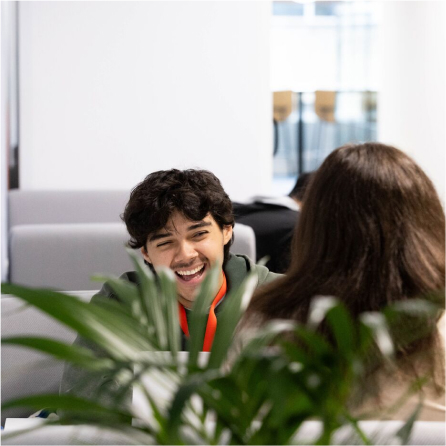 A young man with curly hair and a mustache laughs while talking to a person with long brown hair in a bright room with modern furniture. A green plant is in the foreground.