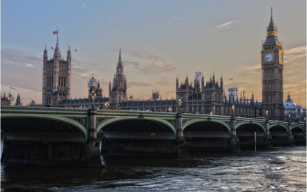 The image shows the Houses of Parliament and Big Ben in London at dusk. The sky is a blend of soft blues and oranges, reflecting off the water of the River Thames in the foreground. The gothic architecture is prominently featured.