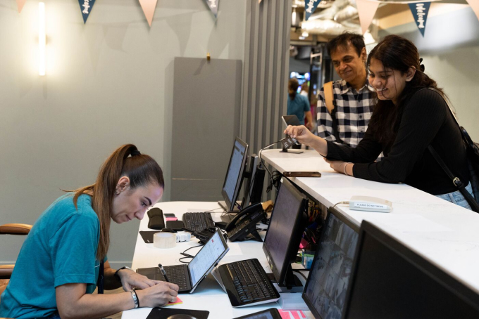 A woman in a teal shirt is sitting at a desk with two laptops, assisting two customers, a man and a woman, who are standing on the other side of the counter. The area is decorated with bunting, and the atmosphere is casual.