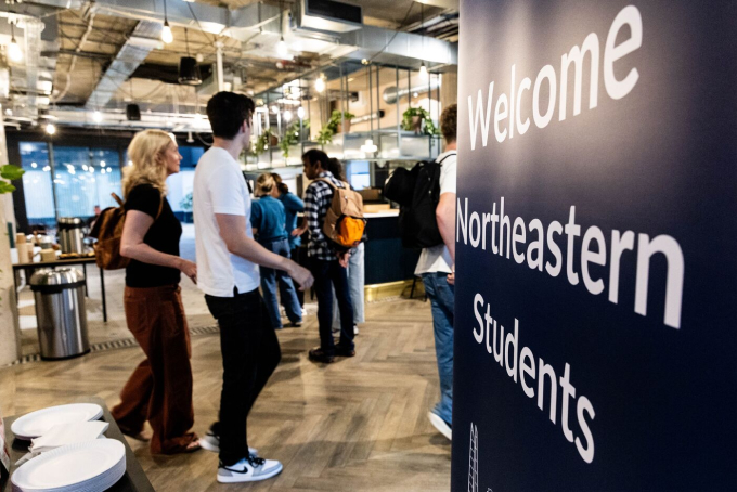 A group of people stands in a modern interior space with exposed ceilings. A large sign in the foreground reads "Welcome Northeastern Students." People appear to be talking and mingling in the background.