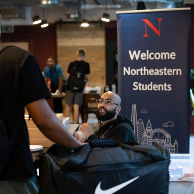 A person sits at a table beside a welcome sign for Northeastern students, engaging with someone carrying a black Nike bag. The room has a casual, welcoming atmosphere with several people in the background.