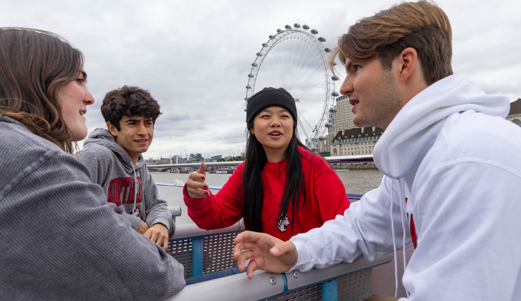 Four people are engaged in conversation on a riverside promenade, with the London Eye visible in the background. The group consists of two men and two women, dressed casually, against a cloudy sky.