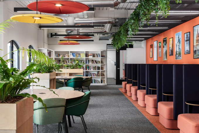 Modern office lounge with colorful decor, featuring round tables, green chairs, and a bookshelf. Bright overhead lights and green plants hang from the ceiling. A row of pink cushioned seats lines an orange wall adorned with framed posters.