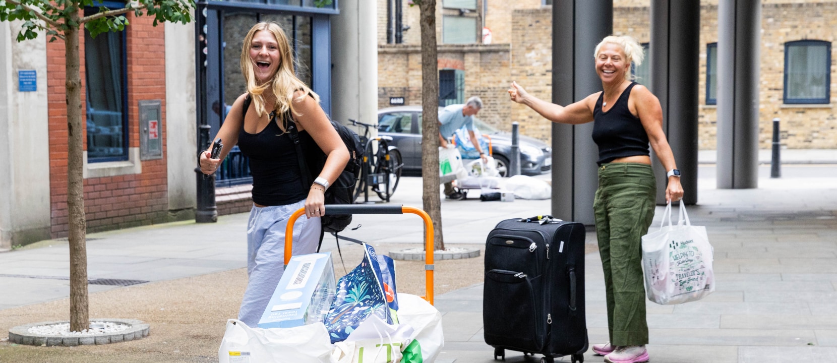 Two women happily wave while carrying bags and pulling a luggage trolley with various items. They stand outdoors on a city street, with brick buildings in the background. One woman wears a black tank top and green pants, the other a black top and gray pants.