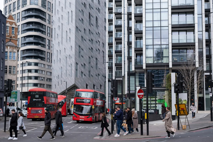 A busy city intersection with pedestrians crossing and two red double-decker buses. Modern high-rise buildings in the background. Overcast sky.