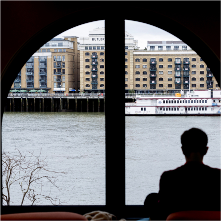 Silhouette of a person sitting by an arched window, overlooking a river and historic buildings across the water. Bare tree branches are visible on the left side outside the window.