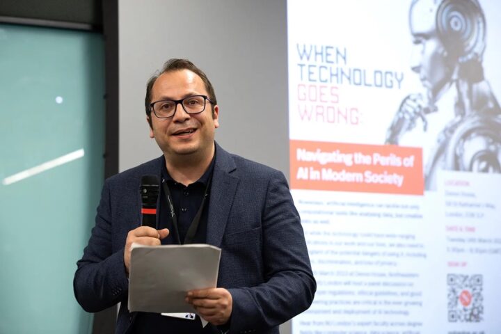 A man holding a microphone and reading from a paper stands in front of a presentation screen displaying the title "When Technology Goes Wrong: Navigating the Perils of AI in Modern Society" with an image of a robot.