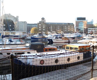 A marina filled with various boats docked in calm waters, surrounded by buildings. A metal chain fence is visible in the foreground, with a clear sky and urban landscape in the background.