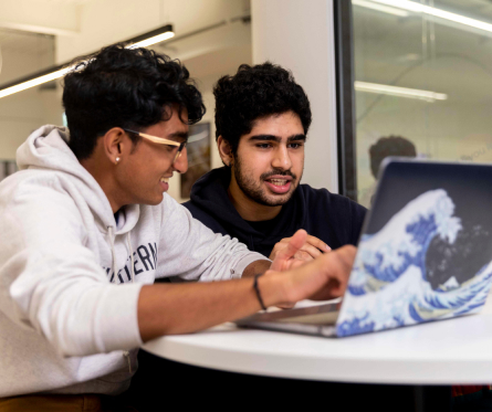 Two people are sitting at a round table, focused on a laptop. One, wearing glasses and a white hoodie, is pointing at the screen, while the other listens intently. The laptop has a wave design on the cover.