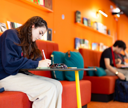 A woman writes in a notebook while sitting on an orange couch in a brightly lit room with books on shelves. Another person is in the background, seated on the same couch, looking down.