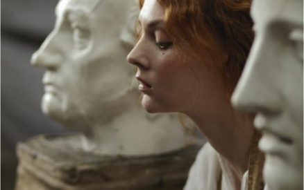 A woman with red hair is leaning in profile between two white stone busts. The focus is on her serene expression as she stands amidst the sculptures.