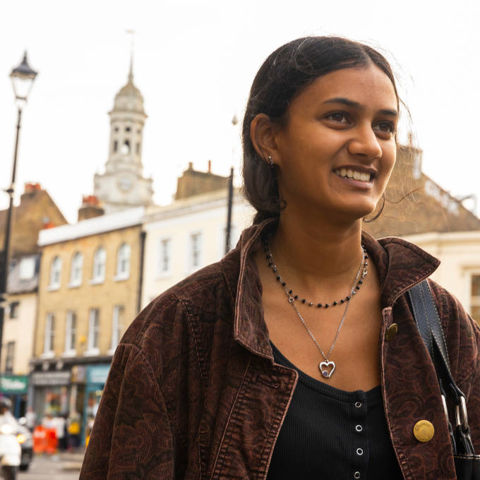 A young woman with long hair smiles, wearing a brown jacket and a heart-shaped necklace. She stands on a street with historic-style buildings in the background, featuring a clock tower and street lamps, on a partly cloudy day.