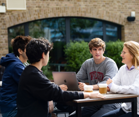 Four people sit at an outdoor table with coffee cups and a laptop, engaged in conversation. The setting is a brick building with large windows and greenery in the background.