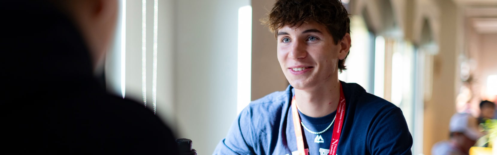 A young person with curly hair and a lanyard smiles while sitting by a window, engaged in conversation. They are wearing a navy blue T-shirt, and sunlight streams through the window, illuminating the room.