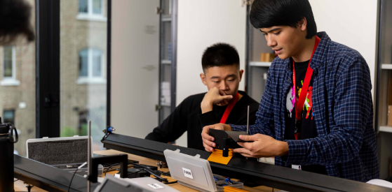 Two people working at a table with electronic equipment. One person is adjusting a device while the other observes thoughtfully. They appear focused and engaged in their task, with a window and building visible in the background.