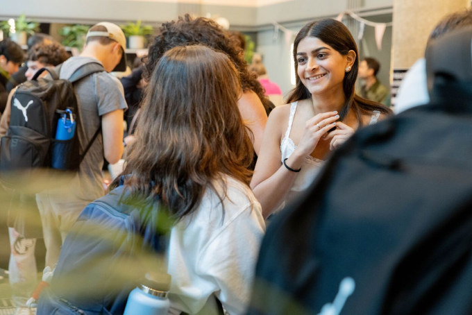 A group of people socializing indoors at a casual event. A smiling woman in a white outfit is engaged in conversation with another person. The room is busy, filled with others wearing backpacks and casual attire.