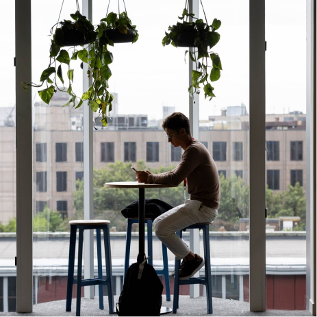 A person sits at a high table using a phone, with hanging plants above and large windows showing a cityscape in the background. Two stools are at the table, and a bag rests on the floor.