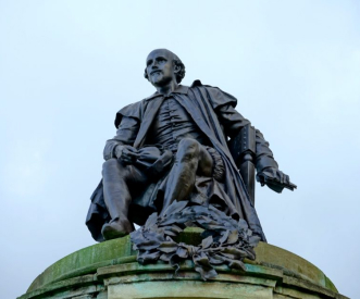 A statue of a man seated on a pedestal, holding a quill in one hand and a scroll in the other. The statue is made of dark metal and captures the figure in period clothing, set against a pale sky background.