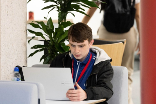 A person with short brown hair is focused on a laptop, sitting in a cozy setting. They wear a jacket and a lanyard. There is a potted plant in the background and other indistinct figures nearby.