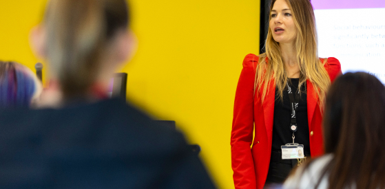 A woman in a red blazer stands in front of a yellow wall, speaking to an audience. She wears a name badge and lanyard. Blurred figures of two audience members are visible in the foreground.