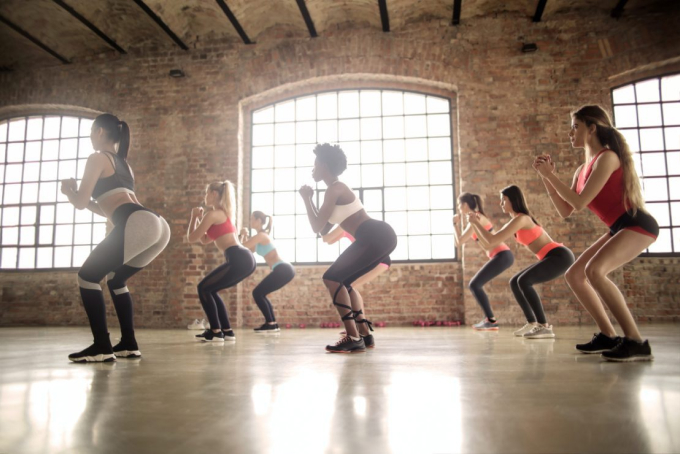 A group of women is exercising in a spacious studio with large, arched windows. They are all performing squats, wearing athletic wear, and appear focused and synchronized. The studio has brick walls and a polished floor.