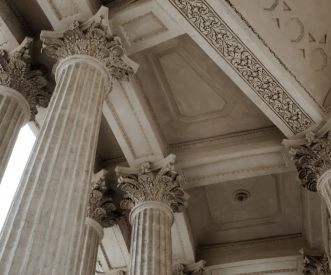 A close-up view of ornate classical columns with intricate capitals and decorative moldings on a ceiling. The architectural details highlight an ancient or neoclassical style.