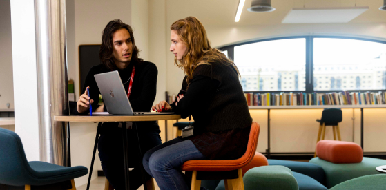 Two people sit at a small round table in a modern office setting. One is typing on a laptop, while the other listens intently. Shelves with books and large windows are in the background.