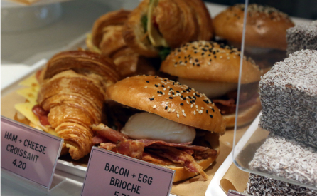 A display of pastries and sandwiches, including ham and cheese croissants, bacon and egg brioche buns with sesame seeds, and chocolate-coated lamingtons, each with visible prices.