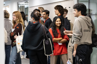 A group of people stands and chats in a corridor next to lockers. Some are leaning against the lockers, and others are facing each other, appearing to be engaged in conversation. Casual attire is visible, and the atmosphere seems relaxed.