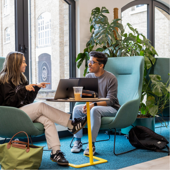 Two people sit in teal armchairs by a window, smiling and talking. A laptop and iced coffee sit on a small table between them. A large green plant is in the background, and backpacks are on the floor.