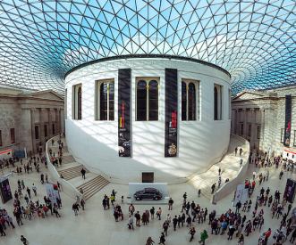 Crowded interior of a museum with a domed glass ceiling, showcasing a large, round white structure adorned with vertical banners. People are walking and gathering on the open floor below, while a classic black taxi is parked nearby.