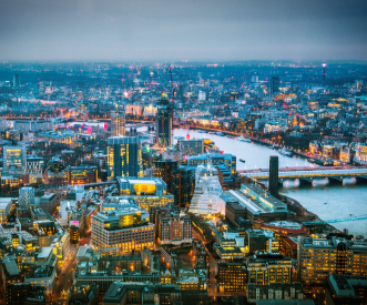 An aerial view of a cityscape at dusk, showcasing illuminated buildings and streets surrounding a river with multiple bridges. The lights create a vibrant atmosphere against the dimming sky.