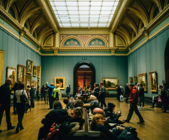 A crowded art gallery with people viewing paintings on the walls. Some visitors are seated on benches in the center of the room. The high ceiling has an intricate design and large windows that allow natural light to illuminate the space.