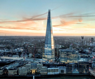 A tall, pointed skyscraper with glass windows reflects the sunset in a cityscape. Surrounding buildings are bathed in warm, colorful light, and the sky features soft, streaky clouds above the urban landscape.