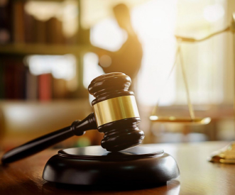 A close-up of a judge's gavel resting on a wooden desk with a blurred scale of justice in the background. A silhouetted person stands near shelves, adding a sense of depth and authority to the setting, illuminated by warm light.