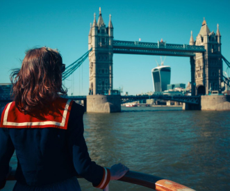 A person wearing a navy and red outfit looks towards Tower Bridge in London from a boat on the River Thames. The sky is clear, and the iconic Walkie Talkie building is visible in the background.