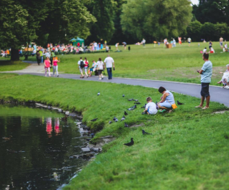 A park scene with people enjoying a sunny day. Children and adults are gathered near a pond, feeding birds. A path curves through the grassy area, and trees are visible in the background.