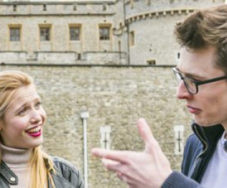 A man and a woman are having a conversation in front of a historic stone castle with arched windows. The woman has blonde hair and is smiling, while the man wears glasses and makes a hand gesture.