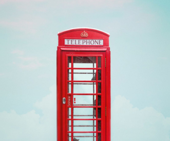 A classic red British telephone box stands against a pastel blue sky with soft clouds. The iconic design features glass panels and a crown symbol on top.