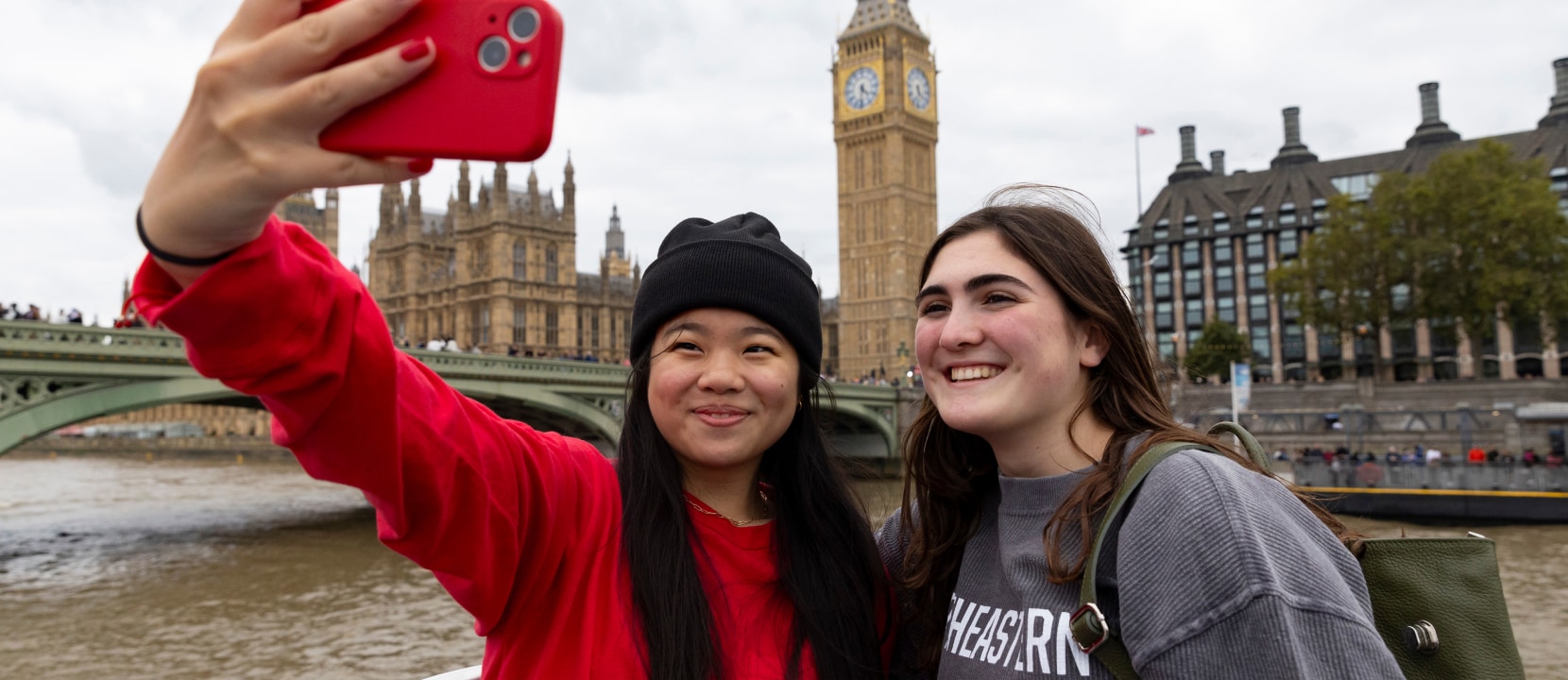 Two people taking a selfie in front of Big Ben and the Houses of Parliament in London. One person wears a red sweater and black beanie, the other a gray sweater. The river Thames and Westminster Bridge are visible in the background.