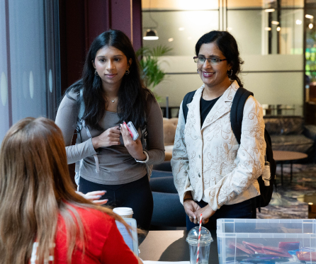 Two women standing and speaking to a seated person at a table indoors. One woman holds a phone and wears a gray top, the other wears a light-colored jacket and glasses. The seated person wears a red shirt.