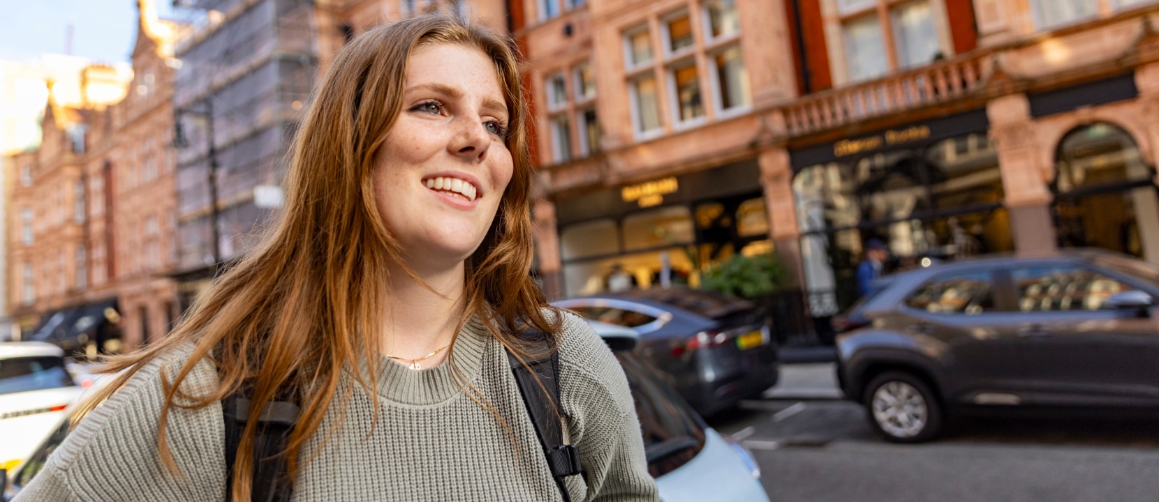 A woman with long hair smiling, wearing a green sweater and backpack, stands on a busy street with parked cars and ornate brick buildings in the background.