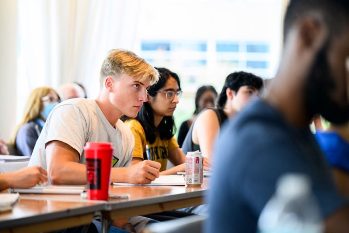 Students sitting at desks attentively taking notes in a classroom. A prominent red water bottle is on one desk. The room is brightly lit and the atmosphere appears focused and engaged.