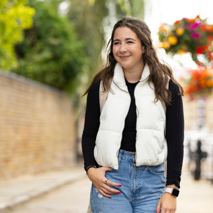 A woman with long brown hair walks outside, wearing a white puffer vest over a black shirt and denim jeans. She smiles while strolling along a path with lush greenery and colorful flowers in the background.