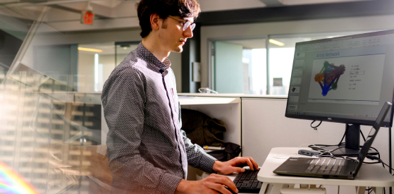 A person wearing glasses and a patterned shirt works at a standing desk. They are focused on a computer screen displaying a presentation with a colorful diagram. The background shows an office setting with large windows.