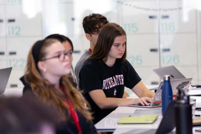 Students seated at desks in a classroom, focused on their laptops. The background shows lockers with numbers. One student in the foreground has long hair and is wearing a black t-shirt. Another student wears glasses and a headband.