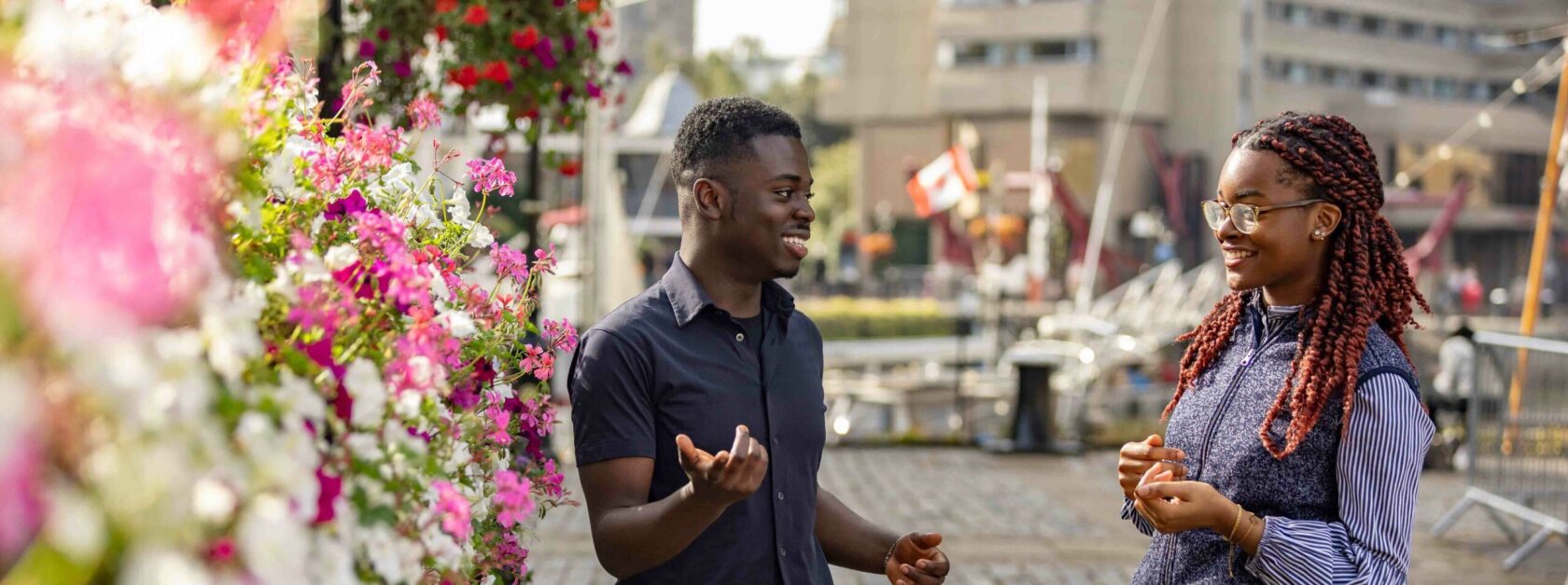 A man and woman smile and talk near a colorful flower display. They stand on a paved area with modern buildings and a flag in the background. The atmosphere is relaxed and pleasant.